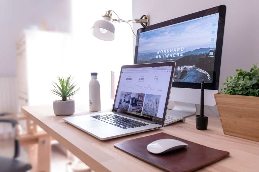 A neatly organized desk with a laptop, desktop computer, mouse, keyboard, potted plant, water bottle, and desk lamp. The desktop screen displays the phrase WORK HARD ANYWHERE over a scenic background.