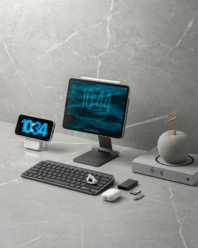 A modern workspace with a wireless keyboard, an iPad on a stand displaying the time, a smartphone dock, wireless mouse, SD cards, AirPods, and a stylish pen holder—a setup perfect for researching why WordPress websites break.