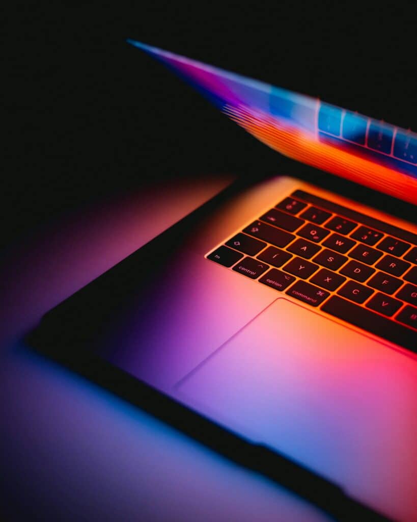A partially open laptop glows with colorful, vibrant light reflecting on its keyboard and surface, creating a dramatic, modern look against a dark background.
