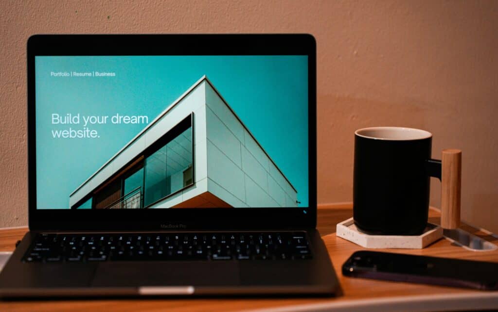 A laptop on a wooden desk displays a website with the text Build your dream website next to a modern building photo, highlighting a care plan for your WordPress website. A black mug, phone, and pen holder sit beside the laptop.
