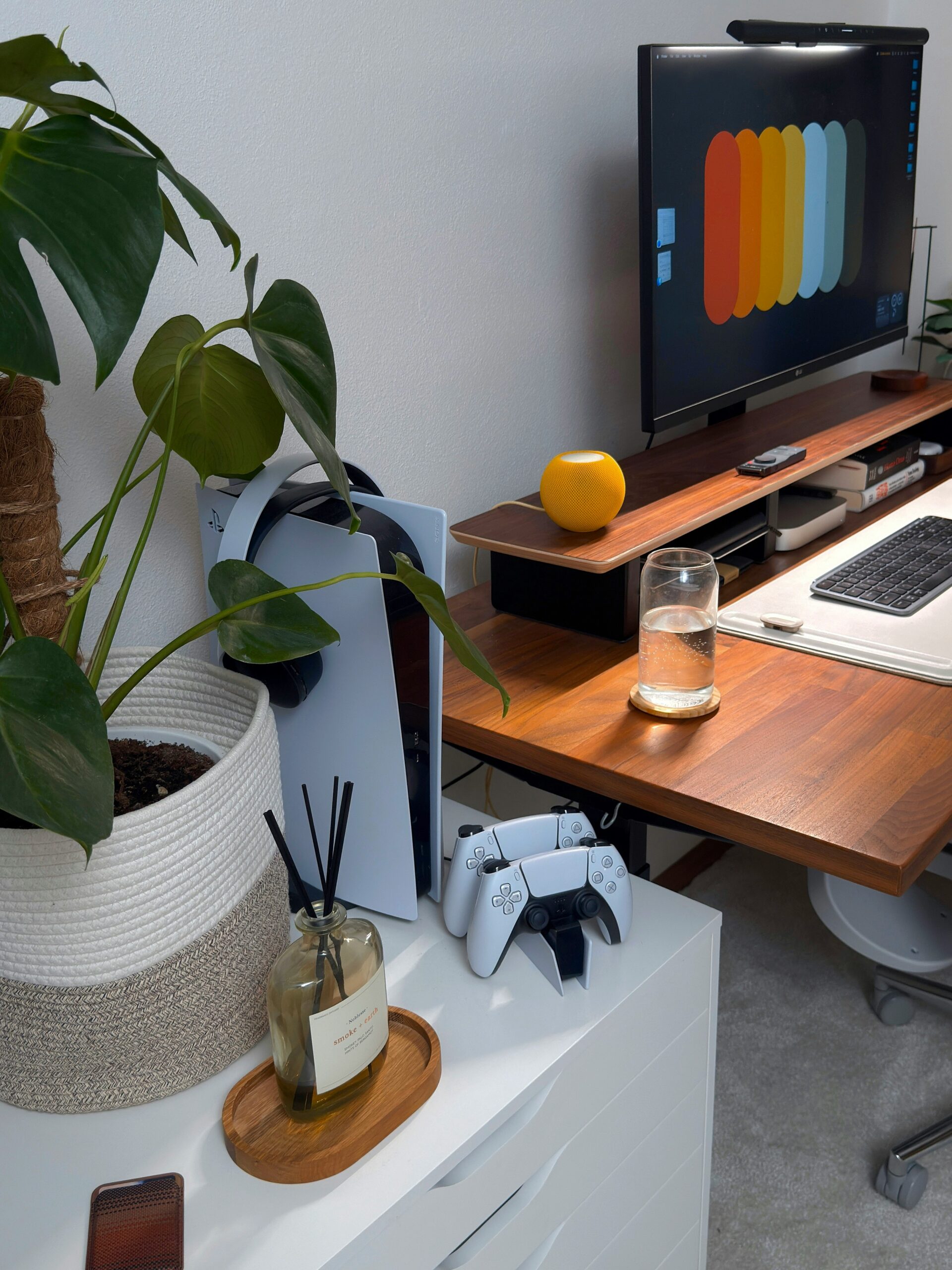 A modern desk setup with a wooden desk, computer monitor displaying color swatches—perfect for reflecting on those times when a WordPress website is over-engineered. PlayStation 5 console, controller, glass of water, candle, potted plant, and reed diffuser complete the scene.