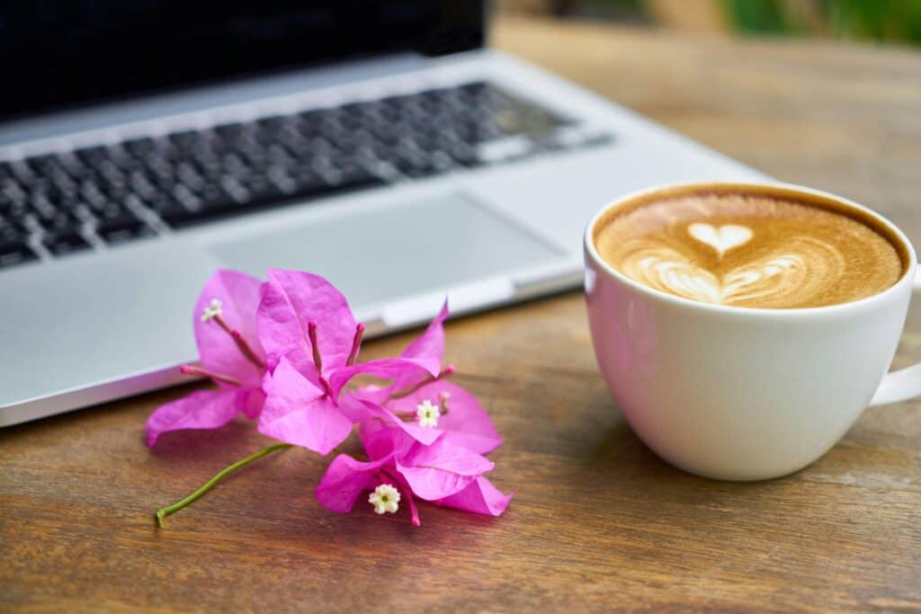 A cup of cappuccino with heart-shaped latte art sits on a wooden table next to pink bougainvillea flowers and a partially visible laptop keyboard, subtly suggesting that a website is a conversation not a brochure—a cozy setting for meaningful digital interaction.